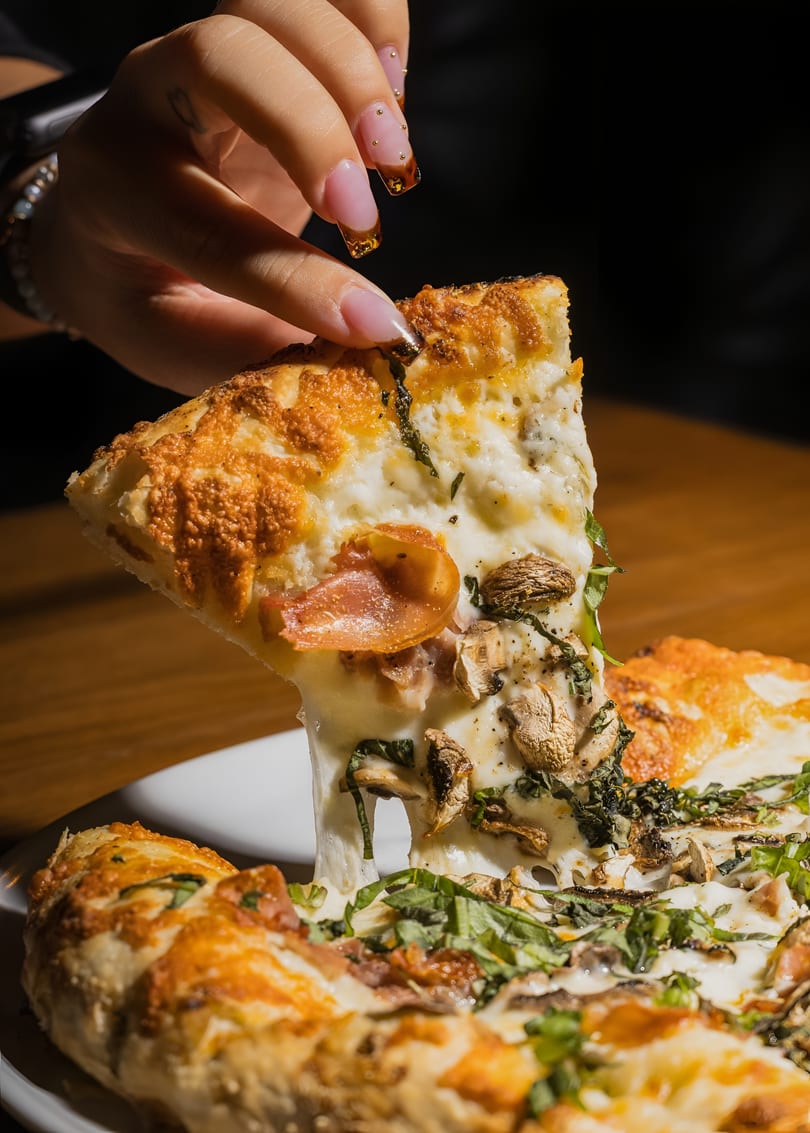 A hand lifts a slice of pizza with melted cheese, mushrooms, prosciutto, and basil.