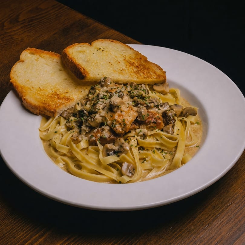 Creamy mushroom and chicken fettuccine pasta with two slices of garlic bread on a white plate.