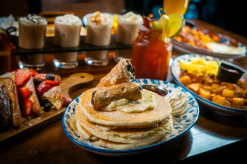 Pancakes with cannolis, French toast, breakfast potatoes, scrambled eggs, and coffee flights on a table.