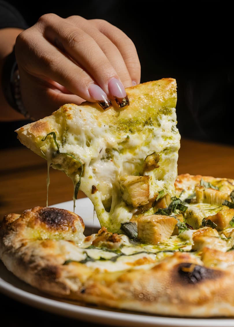 A hand lifts a slice of pesto chicken pizza, with cheese and pesto stretching from the pie.