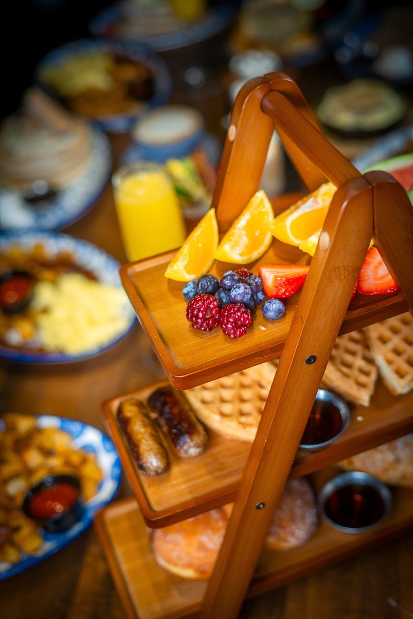 A three-tiered wooden stand holds fresh fruit, waffles, sausages, and donuts, with other breakfast items blurred in the background.