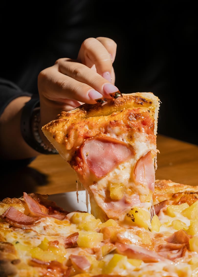 A hand lifts a slice of Hawaiian pizza with melted cheese, ham, and pineapple.