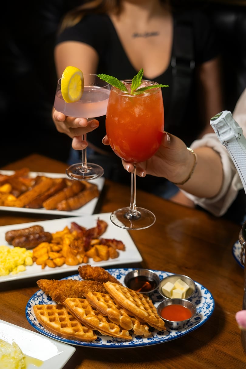 Two people toasting colorful cocktails over a table laden with breakfast dishes like waffles and bacon.