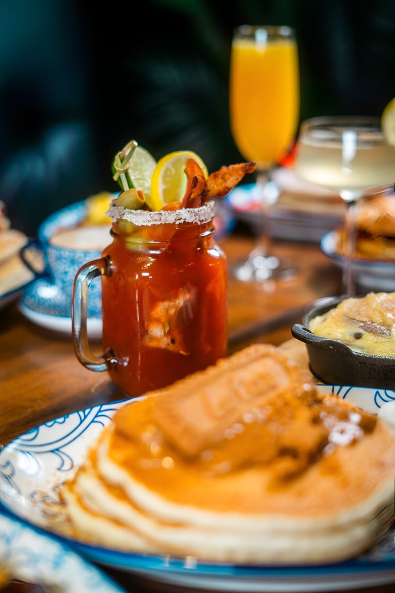 A Bloody Mary with bacon and citrus, pancakes, and other brunch items on a wooden table.